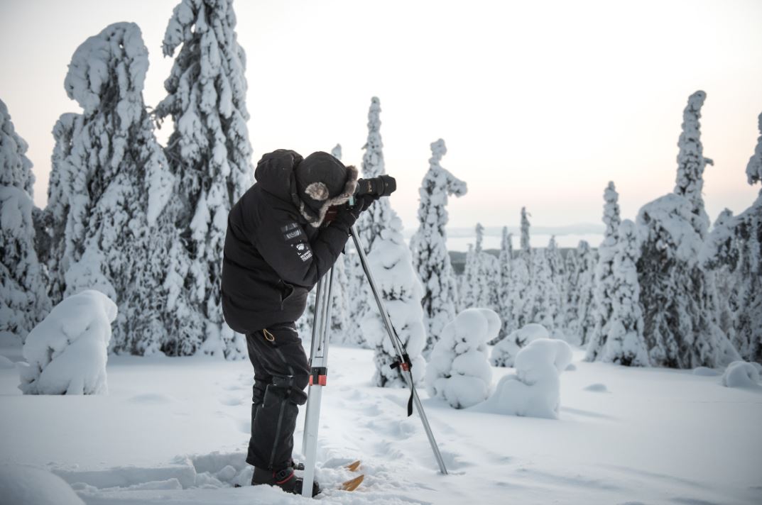Valokuvaaja kameran ja jalustan kanssa talvisessa metsässä.