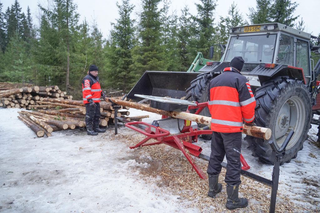 Kaksi miestä kuorii männyn kaarnaa laitteella.