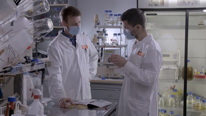 Kaksi miestä valkoisissa takeissa keskustelemassa laboratoriossa. 