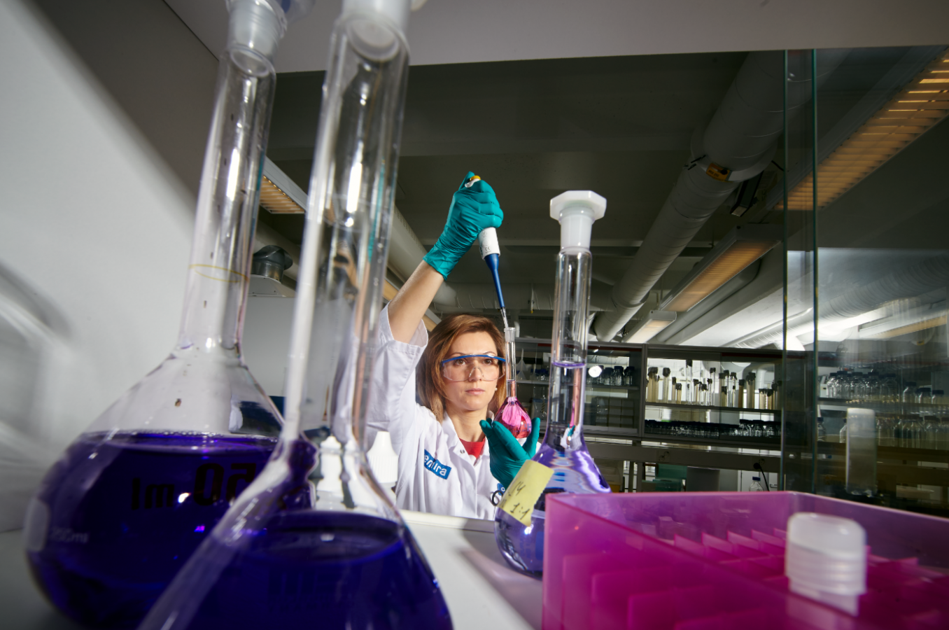 Nainen laboratoriossa pitelee kädessään suurta annostelijaa ja pulloa, jossa on vaaleanpunaista nestettä. Naisella on valkoinen laboratoriotakki, läpinäkyvät suojalasit ja kädessään turkoosit kumihanskat.