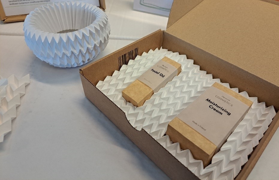 Pöydällä paperista origamitaiteltu pyöreä suojapakkaus ja avonainen pahvilaatikko, jonka sisällä pakkauksissaan ihonhoitotuotteita ja origamitaiteltua paperia. 