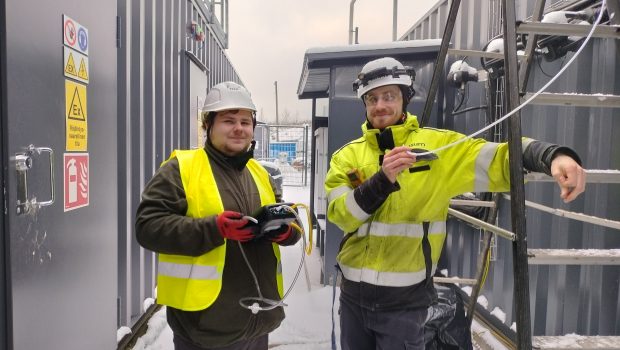 Kaksi mieshenkilöä seisoo turvakypärät ja huomioliivita päällä konttien välissä. Toinen miehistä nojaa tikkaisiin.