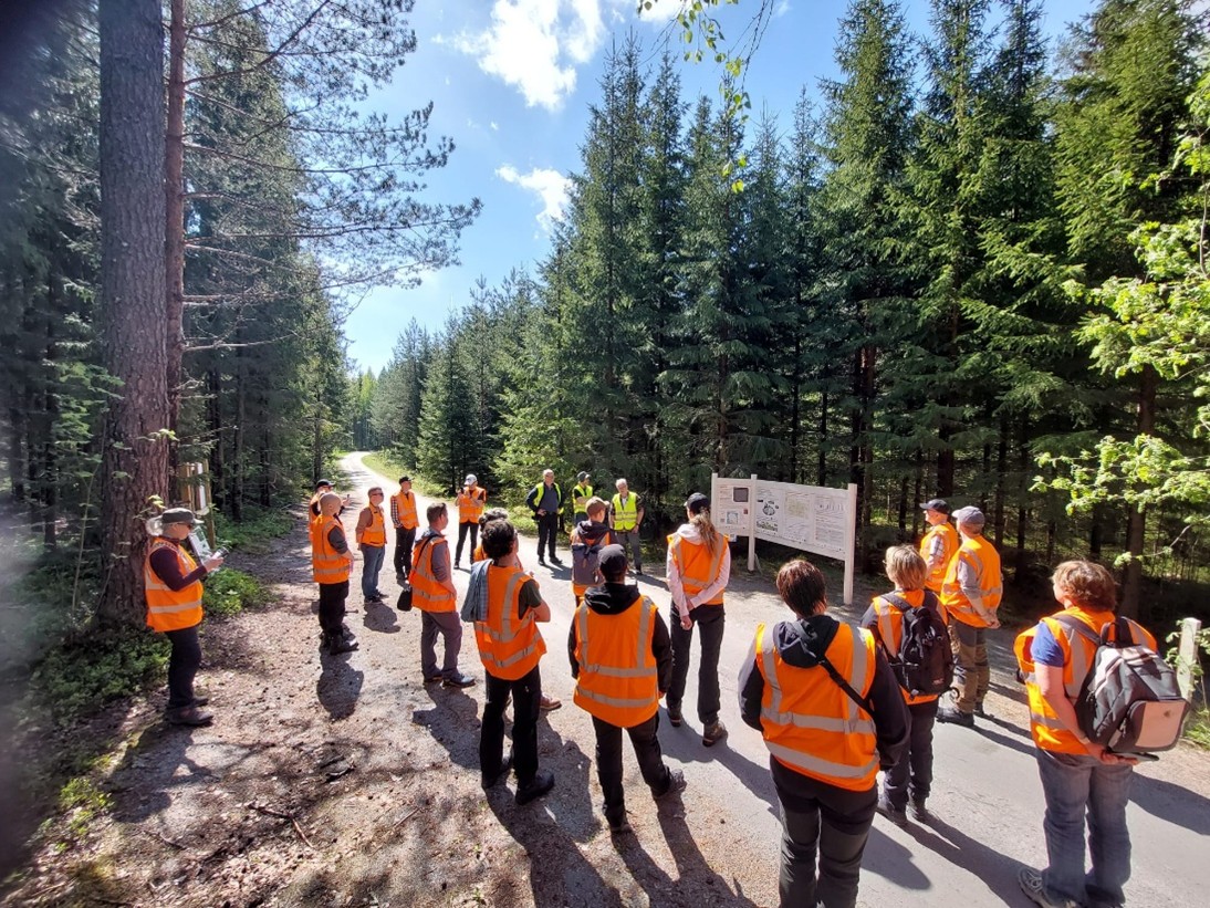 Ihmisryhmä oransseissa huomioliiveissä metsän keskellä kulkevalla polulla infotaulun edessä.
